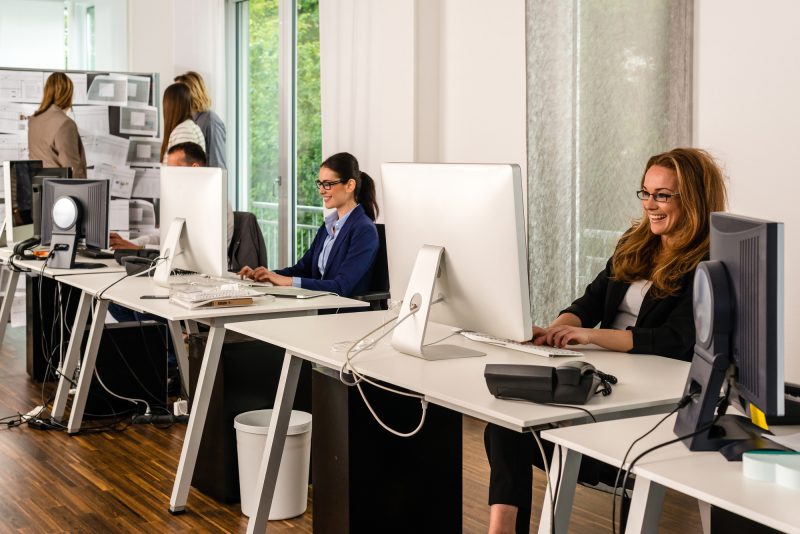 Young team working on computers at desks in stylish office with group discussing conceptual papers in the background, might be a startup or a creative agency